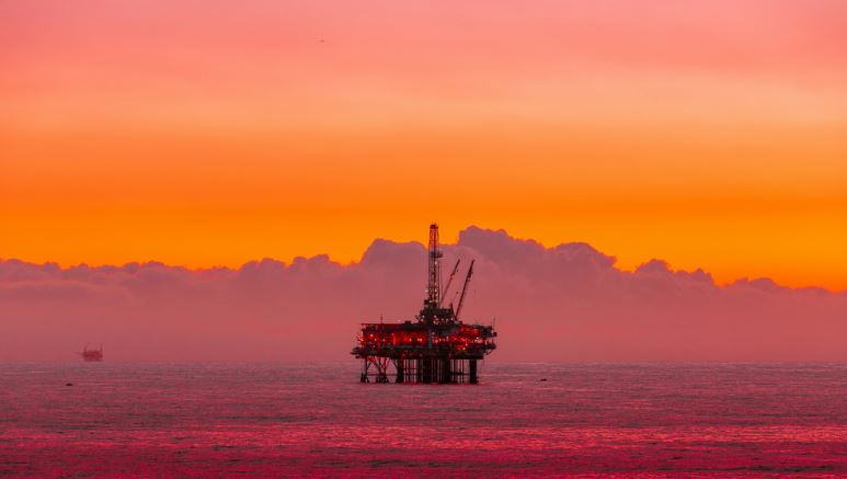 Offshore oil platform illuminated at sunset, silhouetted against a vivid orange sky over calm ocean waters.